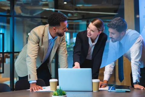 Two men and a woman in professional attire gathered around a table with a laptop, smiling and discussing a business plan.