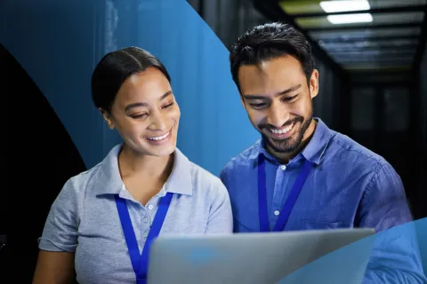 Two professionals wearing ID badges working together on a laptop in a server room, reviewing data and smiling.