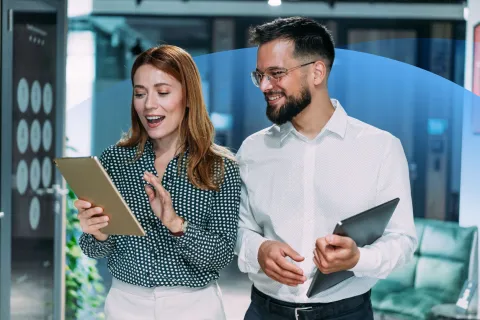 A man and a woman standing together in an office, smiling as they review content on a tablet device.