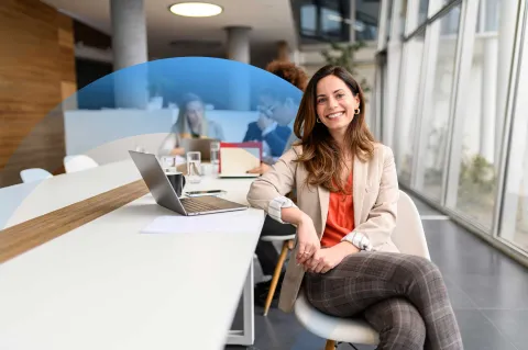 Confident businesswoman sitting at a meeting table, smiling at the camera, with colleagues in the background.