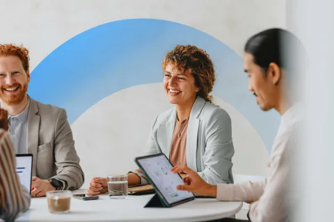 Team members smiling and collaborating during a meeting, using digital tablets and laptops in a bright office.