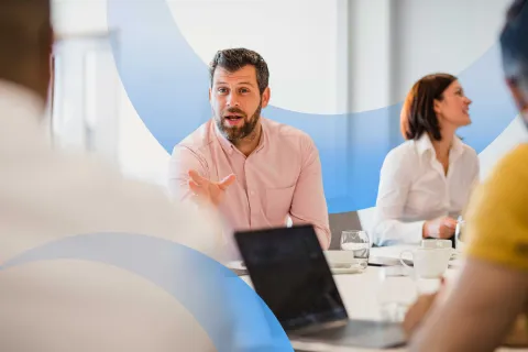 Professional man in a pink shirt leading a meeting discussion with colleagues around a conference table.
