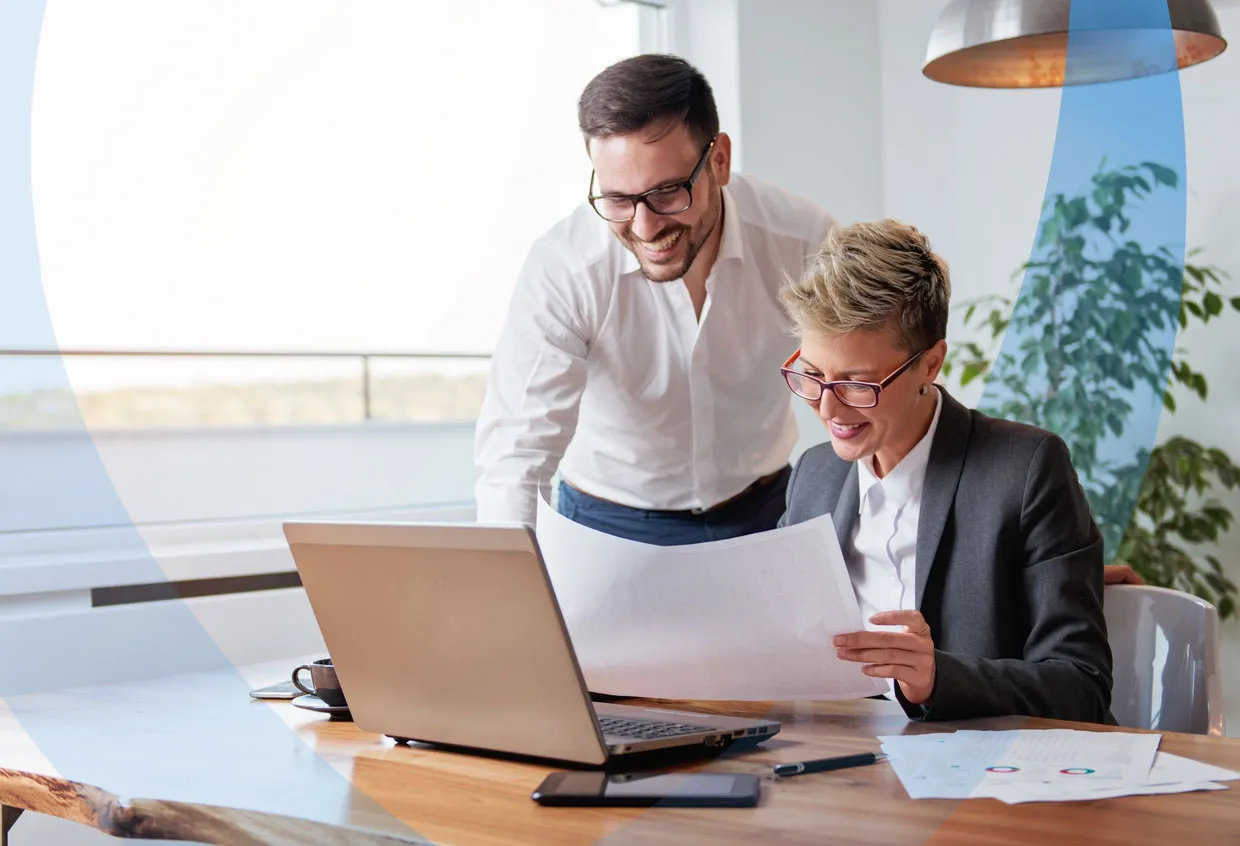 Two business professionals collaborating at a desk, reviewing documents and working together on a laptop.
