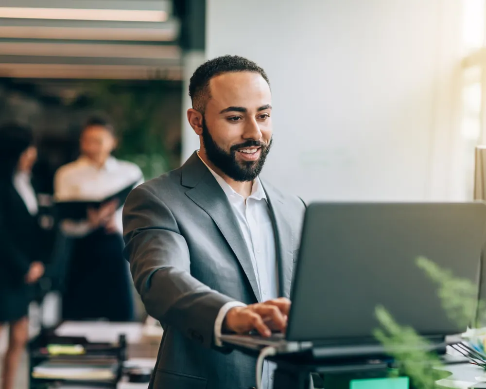Professional in grey suit working on laptop at standing desk in bright office space