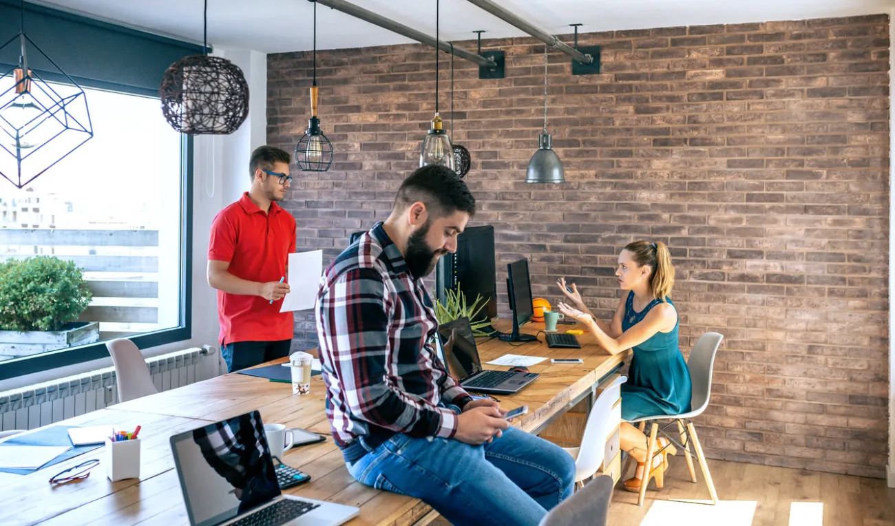 Three people working and chatting in a bright modern office with brick walls