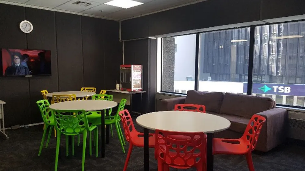 An empty classroom - filled with green and red chairs. Richard Ayoade can be seen on a wall-mounted flat screen tv.