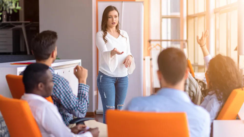 Person in business attire presenting to seated group in orange chairs in sunlit office space