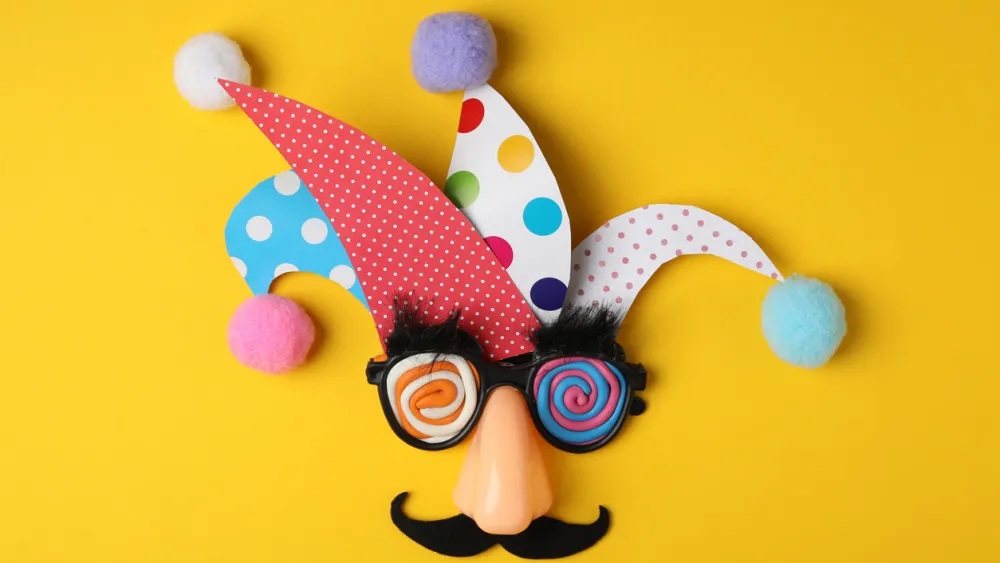 Colorful carnival mask with fake mustache, glasses and pom-poms against a bright yellow background