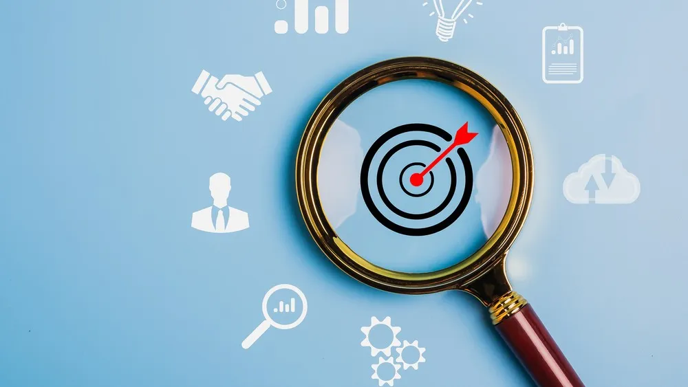 Magnifying glass focused on bullseye target with business icons floating around on blue background