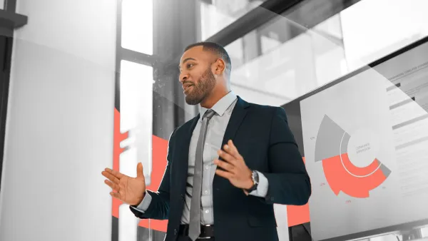 Business professional giving a presentation in an office, explaining data shown on a screen with charts and graphs.