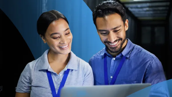 Two professionals wearing ID badges working together on a laptop in a server room, reviewing data and smiling.