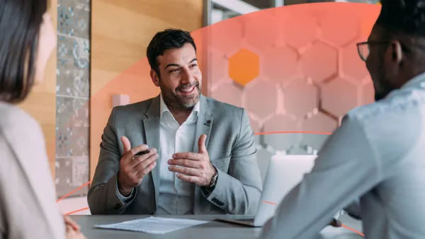 A man in a suit speaking to two colleagues across a table, leading a business discussion.