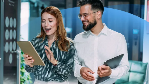 A man and a woman standing together in an office, smiling as they review content on a tablet device.