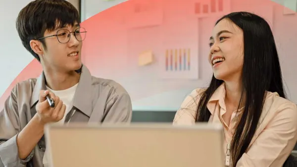 Two colleagues collaborating in an office, smiling and discussing ideas while using a laptop.