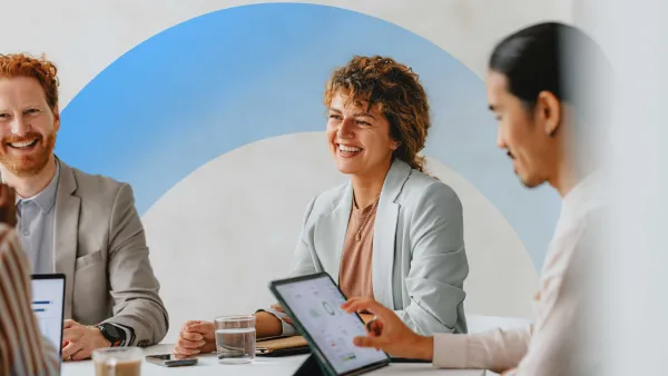 Team members smiling and collaborating during a meeting, using digital tablets and laptops in a bright office.