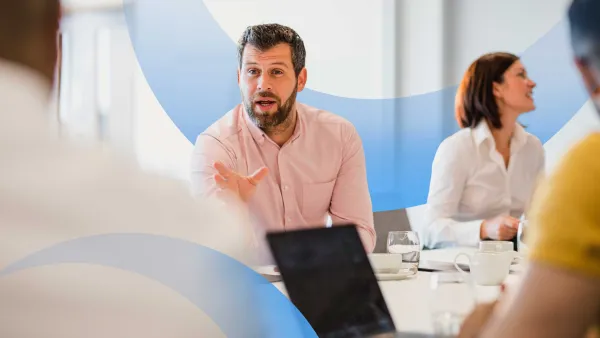 Professional man in a pink shirt leading a meeting discussion with colleagues around a conference table.